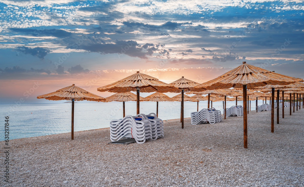 Straw umbrellas with sunbeds on the beach. Straw umbrellas and stacked deck chairs on the beach at sunset or sunrise.