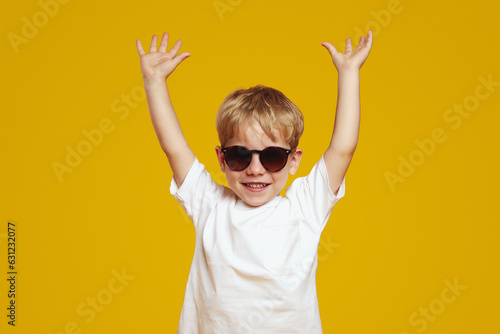 Cheerful little kid with blonde hair, wearing trendy sunglasses smiling and posing with hands up