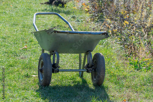 Empty metal wheelbarrow in garden on green grass. Cleaning using gardening equipment in autumn park. Old tool on wheels for garden work.