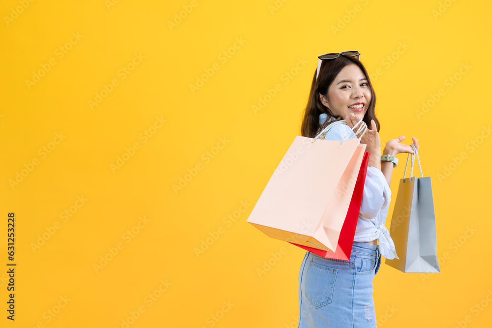 Fototapeta premium portrait of young Asian pretty woman holding shopping bags in hands enjoy buying gifts in store isolated yellow studio background, happy smile shopper spend weekend summer time shopping lifestyle