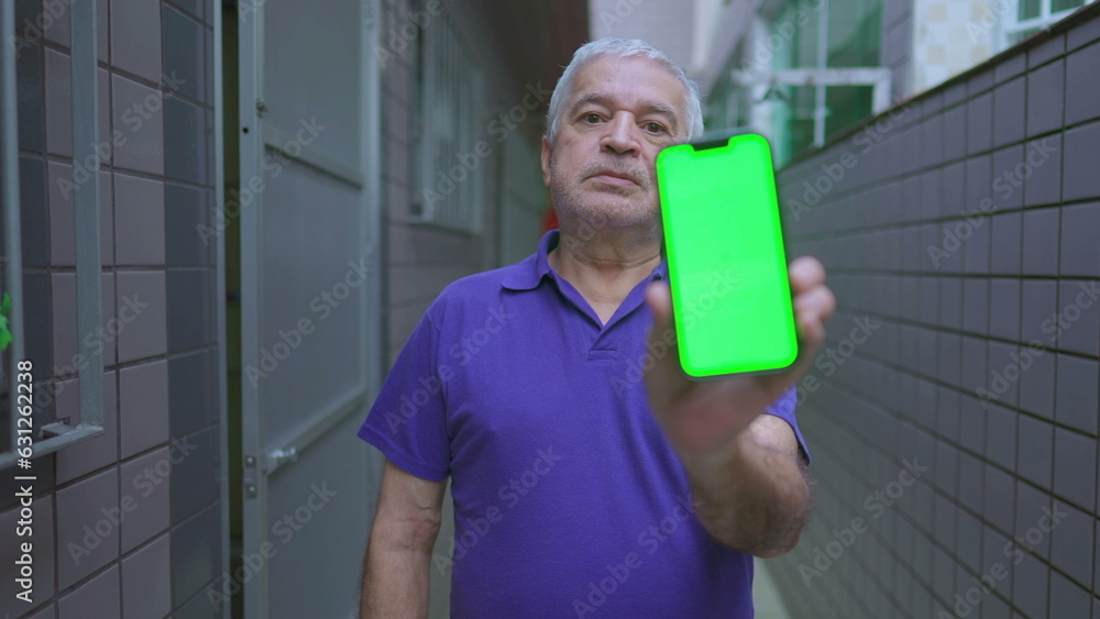 Senior man showing content to camera with chroma key greenscreen on smartphone device, standing in casual domestic residence corridor