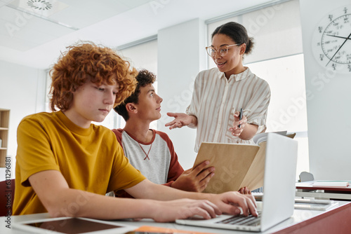 Smiling african american teacher talking to teen schoolboys during lesson with devices in school