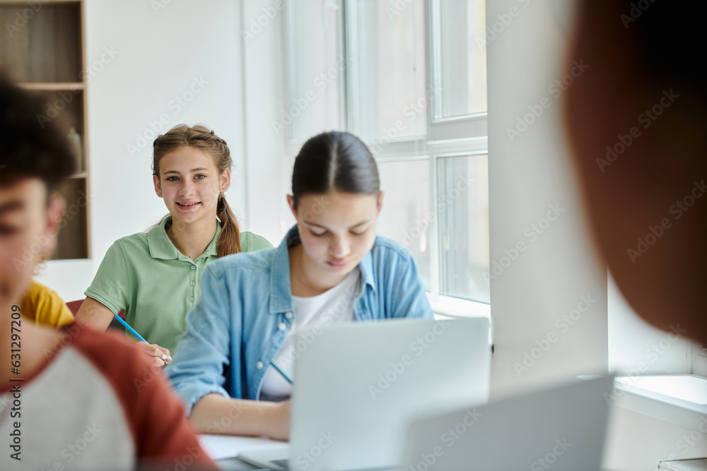 Teen schoolgirl looking at camera and smiling near blurred classmates during lesson in classroom Teen schoolgirl looking at camera and smiling near blurred classmates during lesson in classroom