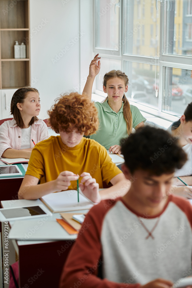 Obraz premium Teenage schoolgirl raising hand and talking near devices and classmates during lesson in school