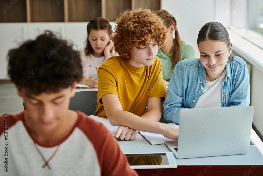 Redhead schoolboy using laptop with smiling classmate together during lesson in blurred school Redhead schoolboy using laptop with smiling classmate together during lesson in blurred school