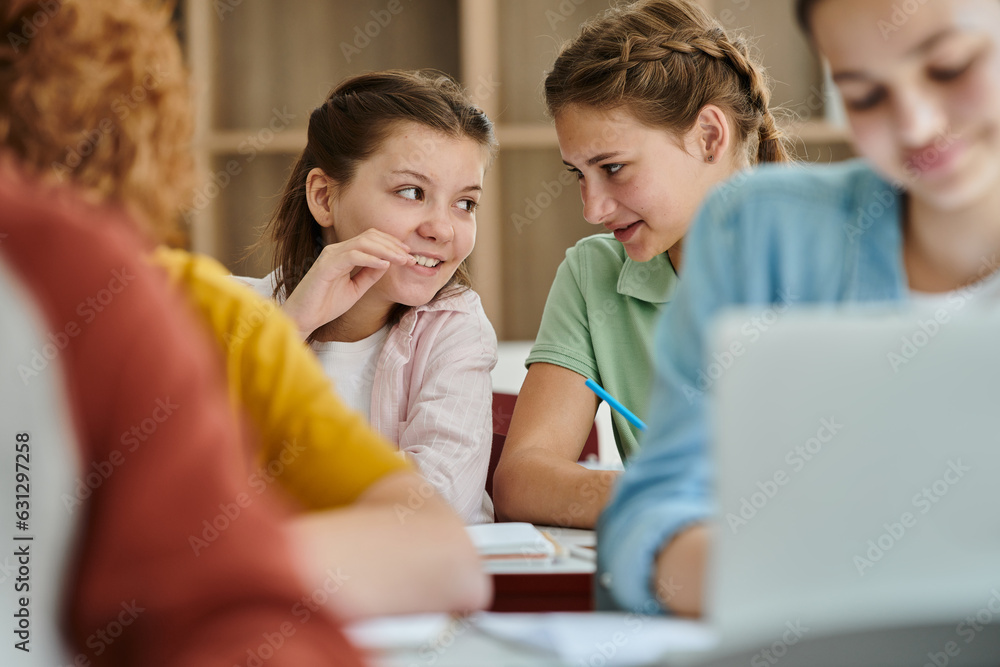 Smiling teenage schoolgirls talking near classmates during lesson in blurred classroom in school Smiling teenage schoolgirls talking near classmates during lesson in blurred classroom in school