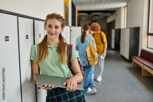 positive teen girl holding laptop and looking at camera in hallway, back to school concept