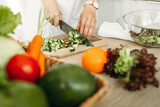 A woman cuts a cucumber with a knife on the background of vegetables lying on the table.