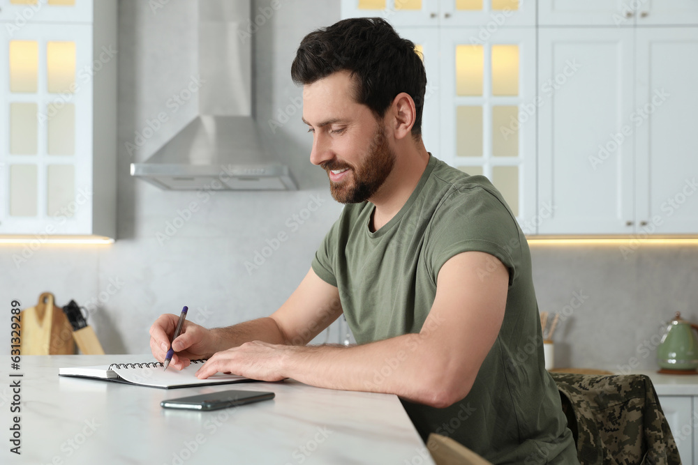 Soldier writing in notebook at white marble table in kitchen. Military service Soldier writing in notebook at white marble table in kitchen. Military service