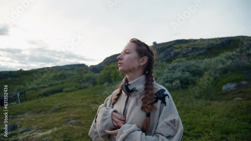 A young caucaisan girl with a thoughtful face, brown hair and pigtails stands, looks up and zips up her light-colored jacket on a rocky cliff and hills in Norway on a cloudy day photo