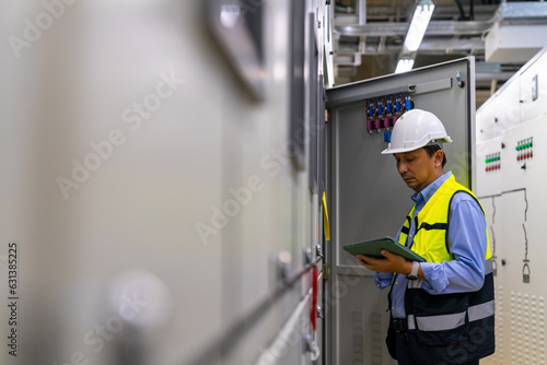 Professional Asian male engineer in safety uniform working at factory server electric control panel room. Industrial technician worker maintenance checking power system at manufacturing plant room.