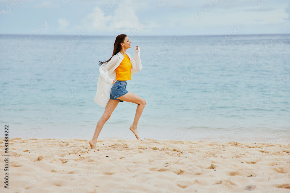 Fototapeta premium Sports woman runs along the beach in summer clothes on the sand in a yellow T-shirt and denim shorts white shirt flying hair ocean view