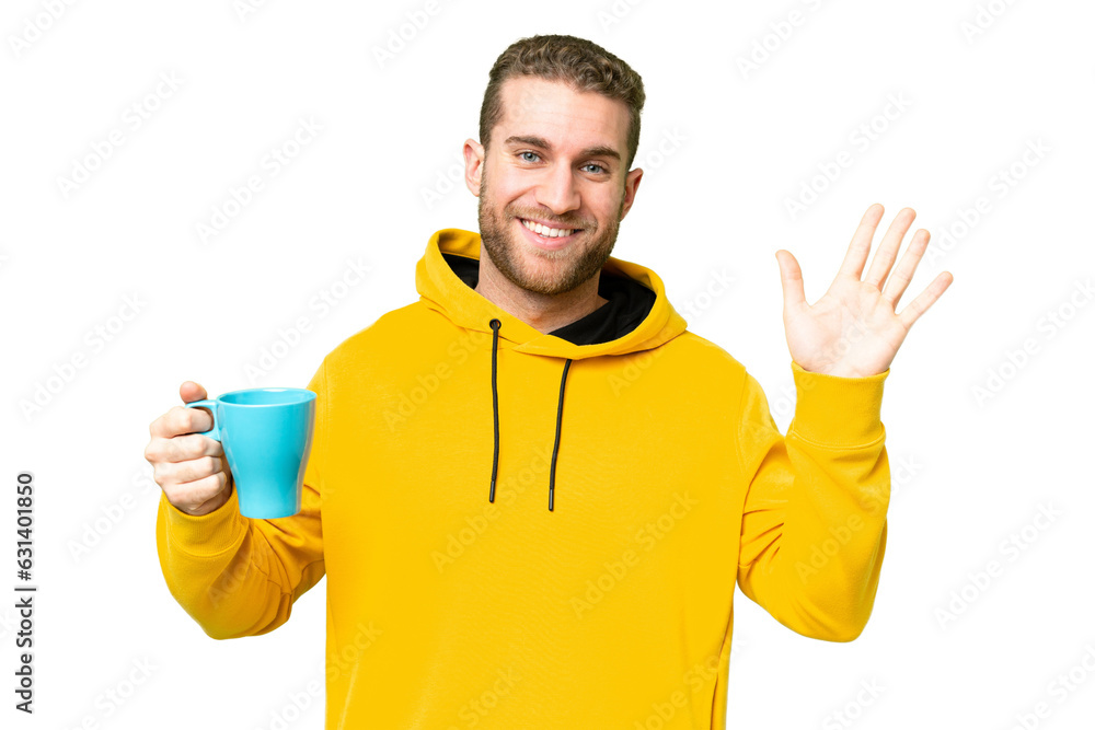 Young handsome blonde man holding cup of coffee over isolated background saluting with hand with happy expression Young handsome blonde man holding cup of coffee over isolated background saluting with hand with happy expression