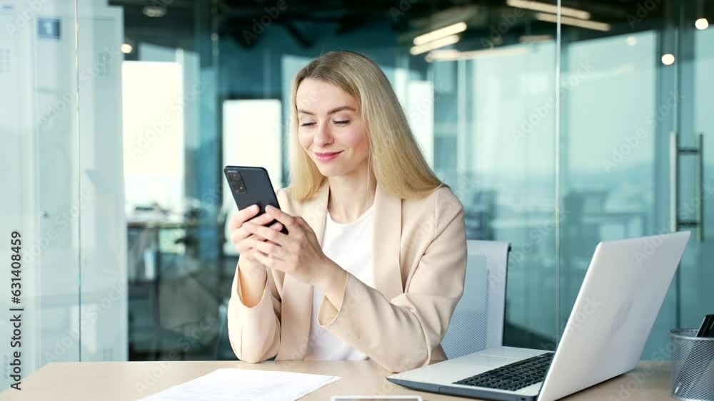 Young happy businesswoman in a suit is using smartphone while sitting at workplace in modern office. Smiling blonde woman shopping online, browsing social media, typing message, chatting with a client Young happy businesswoman in a suit is using smartphone while sitting at workplace in modern office. Smiling blonde woman shopping online, browsing social media, typing message, chatting with a client