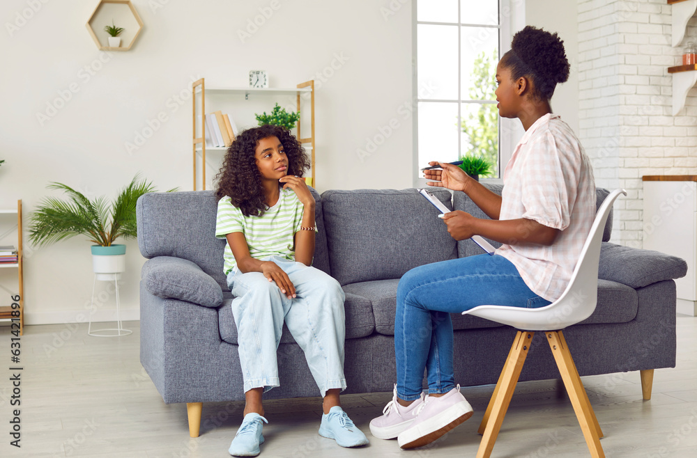 Professional psychologist talking to child. Little African American girl  sitting on sofa and talking to woman therapist or psychologist who is  sitting on chair, holding clipboard and asking questions Stock Photo