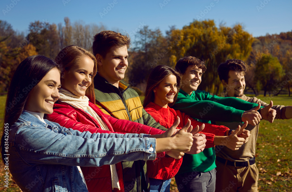 Group of happy young friends showing thumbs up all together. Several cheerful, joyful people standing in a row in a sunny autumn park, looking away, giving thumbs up and smiling Group of happy young friends showing thumbs up all together. Several cheerful, joyful people standing in a row in a sunny autumn park, looking away, giving thumbs up and smiling