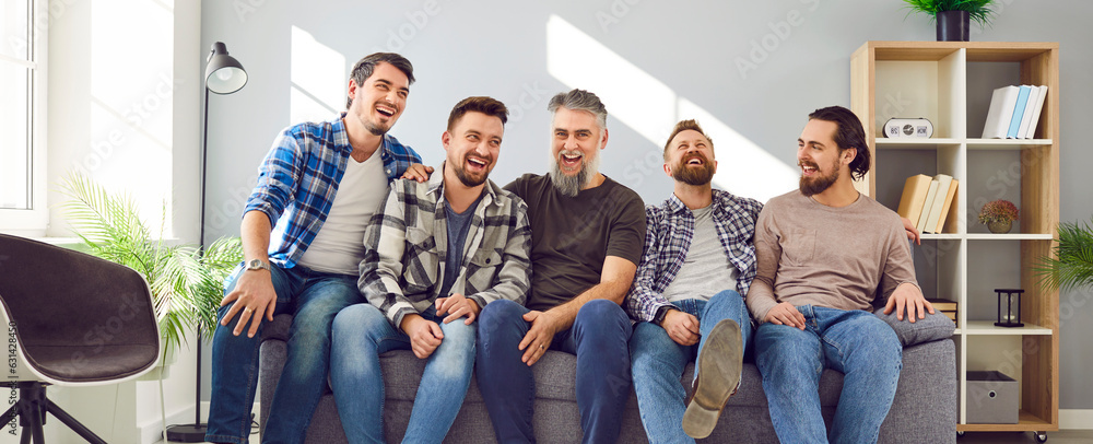 Happy male best friends sitting on couch together. Cheerful hipster men in casual wear laughing and enjoying leisure time while relaxing on sofa in apartment. Meeting of best friends, friendship