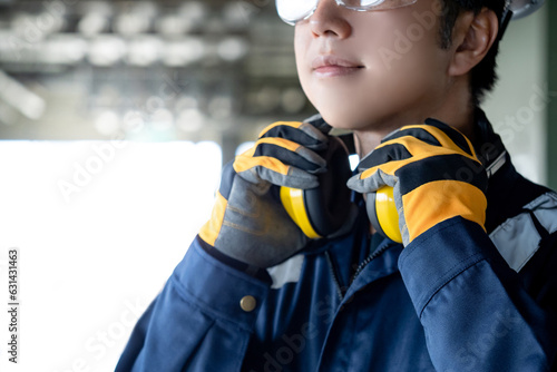 Asian man construction worker wearing uniform suit, safety helmet, goggles and protective gloves holding yellow ear muffs or ear defenders on his neck at construction site