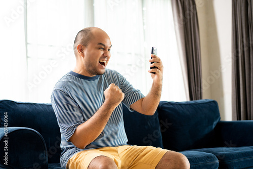 Happy shocked face Young asian man playing game with smartphone sitting on blue sofa in the house. Winner Man with typing cellphone  while sitting on couch in living room