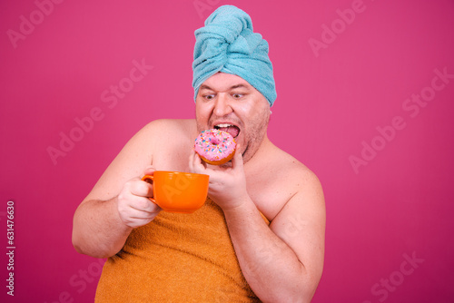 Early morning. Funny fat man drinks coffee and eats donuts. The guy wrapped in a towel after a shower poses on a pink background.