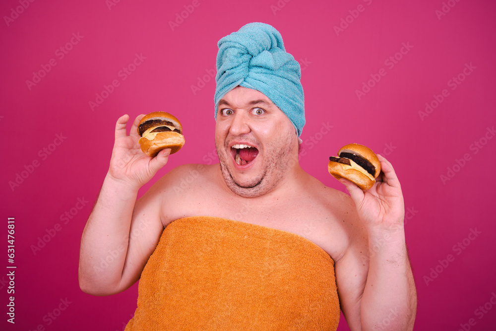 Early morning. Funny hungry fat man eating burgers. The guy wrapped in a towel after a shower poses on a pink background. Early morning. Funny hungry fat man eating burgers. The guy wrapped in a towel after a shower poses on a pink background.