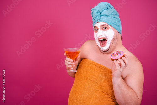 Early morning. Funny fat man drinks a cocktail before a party. The guy wrapped in a towel after a shower poses on a pink background.