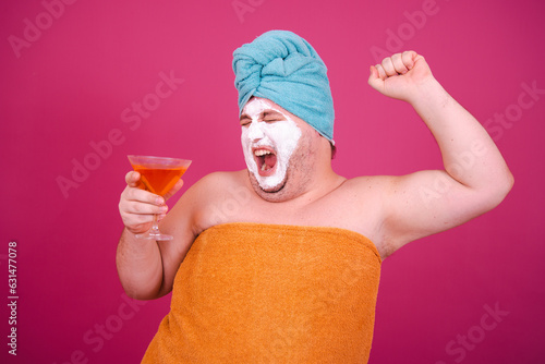Early morning. Funny fat man drinks a cocktail before a party. The guy wrapped in a towel after a shower poses on a pink background.