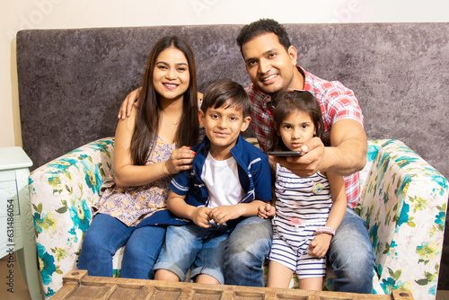 Happy Young indian family sitting on sofa couch at home looking at television. Parents with two kids change channel with TV remote.