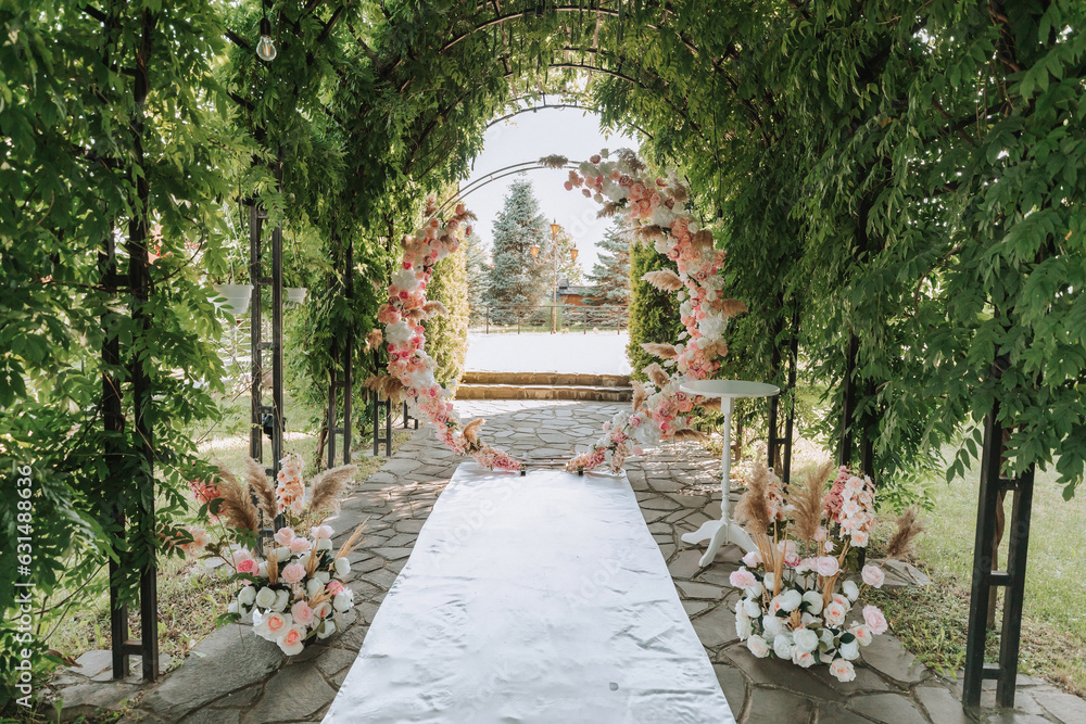 The wedding arch in the park is made of fresh flowers and dry reeds. Outdoor wedding ceremony in nature in a green corridor. White chairs for the ceremony