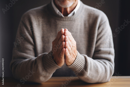 Unrecognizable senior man in gray sweater at home in his living room praying, hands clasped together, eyes closed