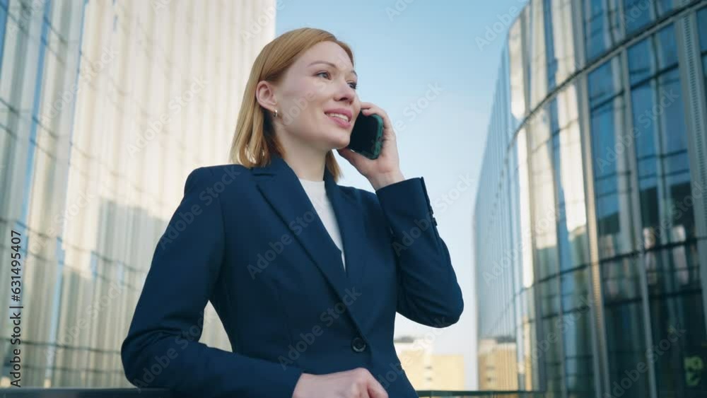 Gimbal shot positive blond hair business woman wearing blue formal suit talking on smartphone and smiles standing on office building rooftop leans on glass railing of terrace in city downtown Gimbal shot positive blond hair business woman wearing blue formal suit talking on smartphone and smiles standing on office building rooftop leans on glass railing of terrace in city downtown