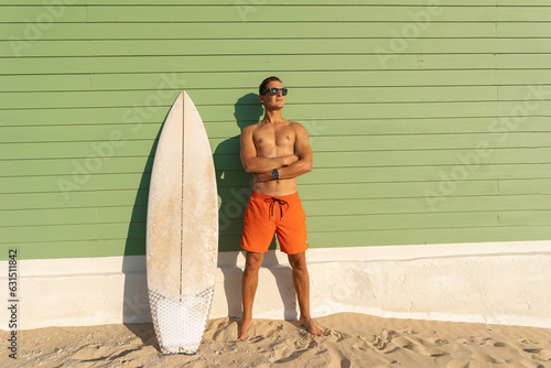 A smiling joyful man with nice body wearing sunglasses standing at the light green with a surfboard
