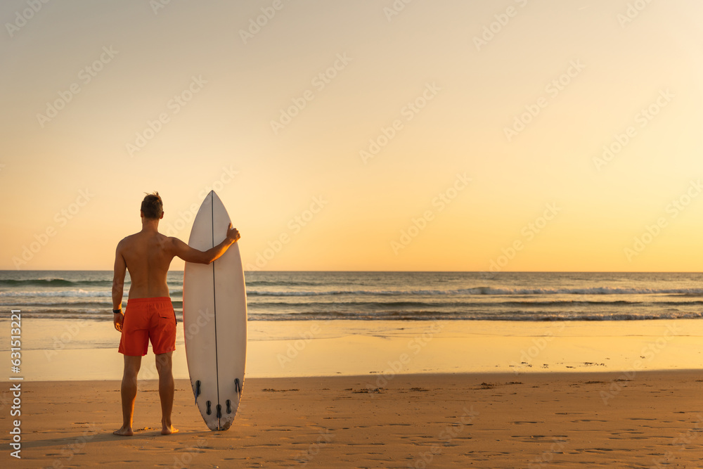 A man standing on the shore holding a surfboard - view from the back