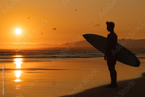 Black silhouette of an athletic man standing on the shore holding a surfboard at orange sunset