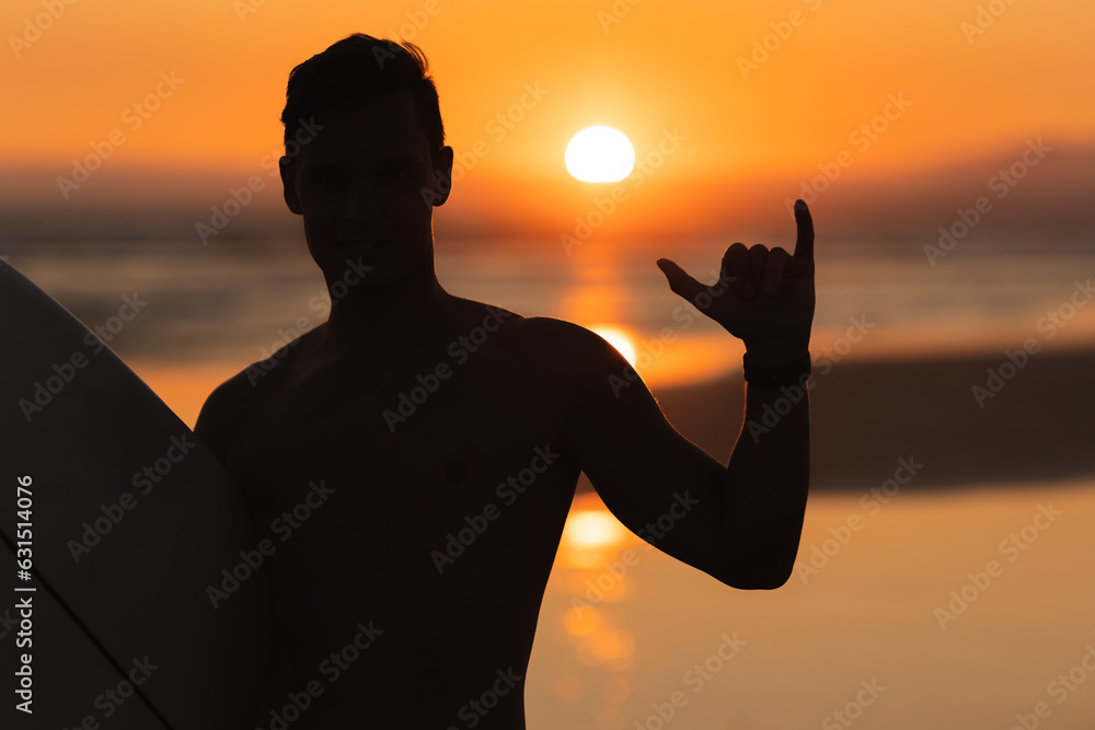 Black silhouette of an athletic man surfer showing shaka at sunset