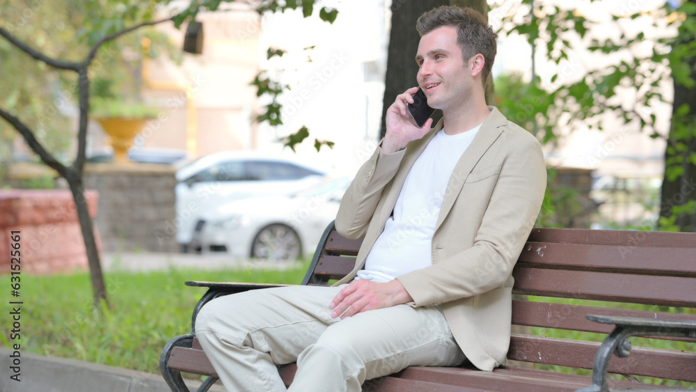 Fototapeta premium Casual Young Man Talking on phone while Sitting Outdoor on a Bench
