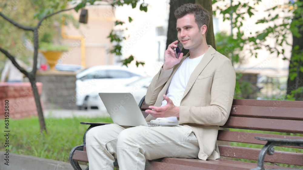Casual Young Man Talking on Phone and Using Laptop Outdoor Casual Young Man Talking on Phone and Using Laptop Outdoor