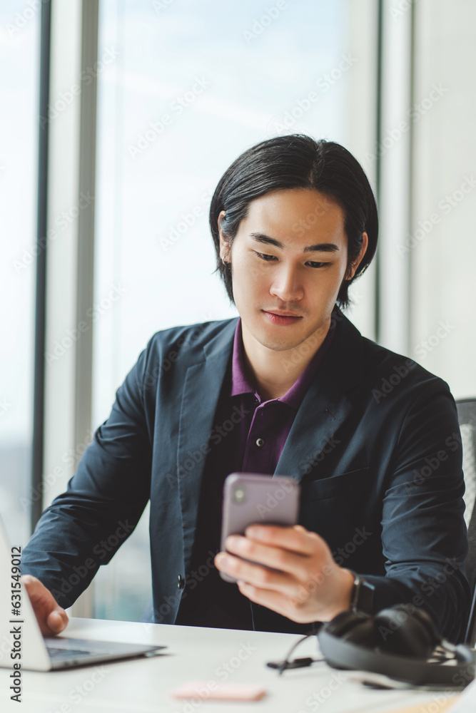 Stylish asian businessman holding mobile phone, sitting at workplace, using laptop in modern office Stylish asian businessman holding mobile phone, sitting at workplace, using laptop in modern office
