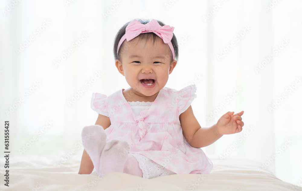 Adorable Asian baby laughing, looking at camera, sitting on bed. Smiling little cute toddler girl wear casual pink dress, headband and socks. Healthy child with happy expression face. White background