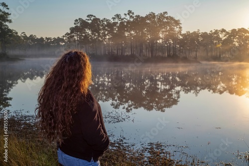 Female with curly hair admiring the lake with vegetation and sunset reflection © Adrian De La Paz/Wirestock Creators