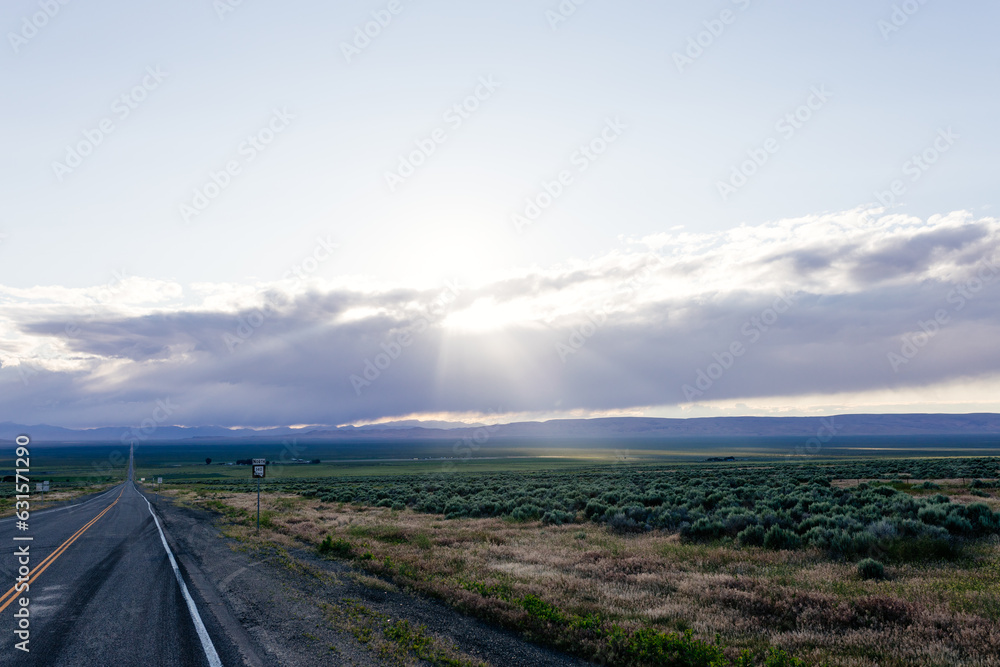 A long asphalted straight road stretches far into the horizon, on which blue mountains can be seen. The rays of the sun break through the large clouds over the road. blue sky before sun A long asphalted straight road stretches far into the horizon, on which blue mountains can be seen. The rays of the sun break through the large clouds over the road. blue sky before sun