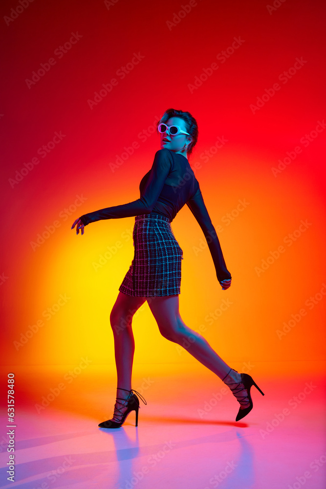 Full lenght photo of running woman, young fashion model wearing fashionable in transparent top and skirt posing over gradient yellow-red neon background.