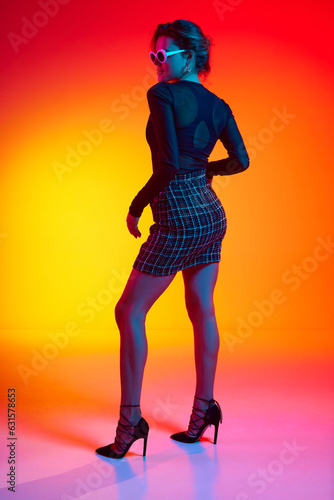 Full lenght photo of woman wearing fashionable with sunglasses in transparent top and skirt posing over gradient yellow-red neon background. Backview