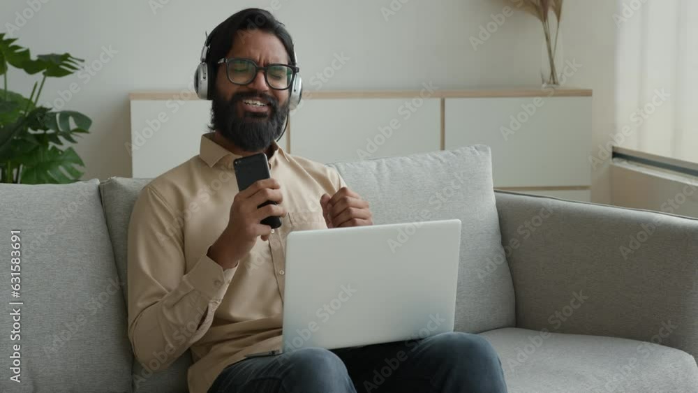 Happy relaxed millennial man Arabian Indian muslim guy on couch cozy sofa listen music in headphones work break lazy weekend listening song audio track use mobile phone working online laptop at home Happy relaxed millennial man Arabian Indian muslim guy on couch cozy sofa listen music in headphones work break lazy weekend listening song audio track use mobile phone working online laptop at home