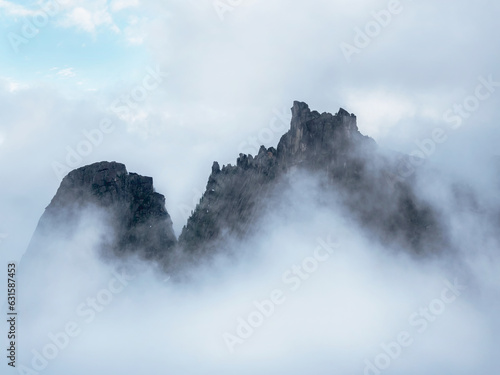 Top mountains in white clouds. The edge of the sheer cliff is barely visible due to low clouds and dense fog. White cloud descended on the red mountain peaks. Light in the morning mountains.