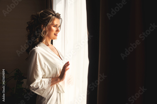 Sensual woman in home bathrobe with nice makeup and hairstyle look at window in living room, happy looking away. Lovely lady posing at home. Concept of luxury fashionable style. Copy ad text space