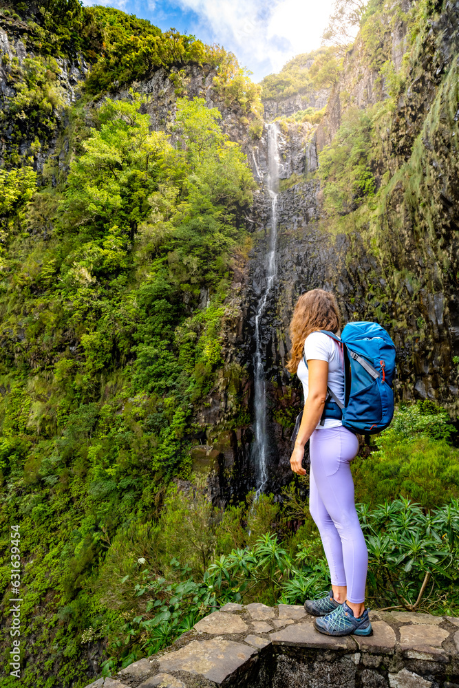 Female tourist with backpack standing on stone wall viewpoint and watches green overgrown Risco waterfall. 25 Fontes Waterfalls, Madeira Island, Portugal, Europe.