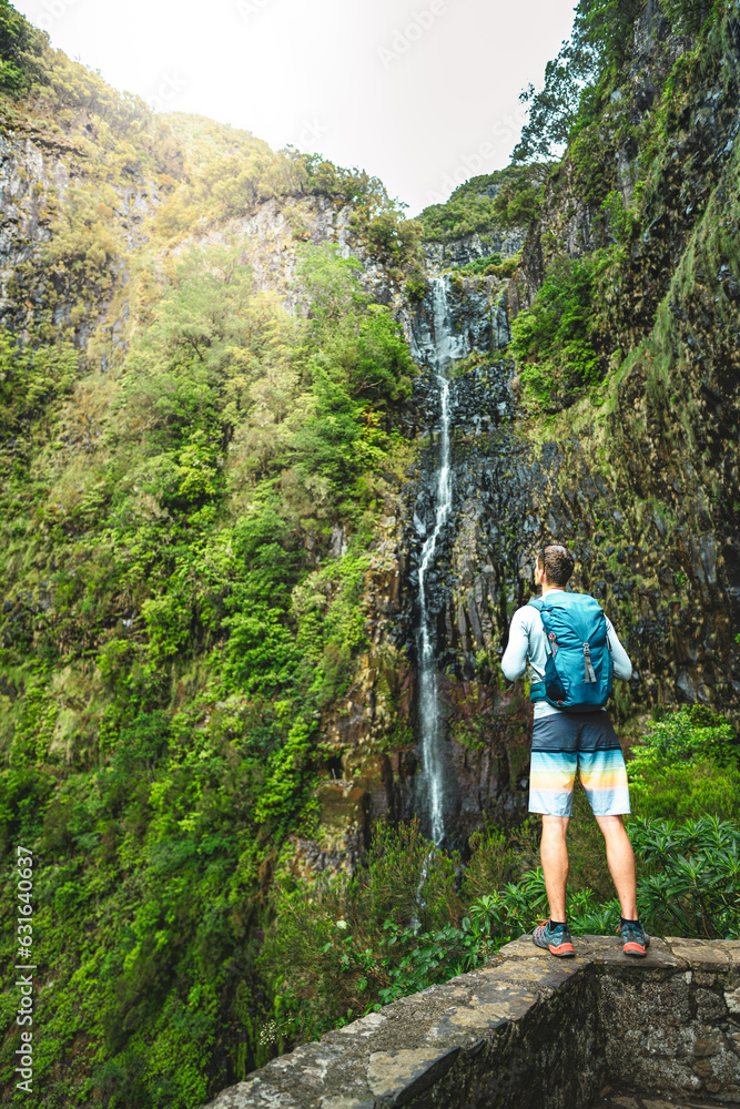 Tourist with backpack standing on stone wall viewpoint and watches green overgrown Risco waterfall. 25 Fontes Waterfalls, Madeira Island, Portugal, Europe. Tourist with backpack standing on stone wall viewpoint and watches green overgrown Risco waterfall. 25 Fontes Waterfalls, Madeira Island, Portugal, Europe.