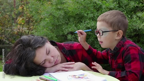 A young tired mother is sleeping, the child is trying to wake her up. The child has vision problems, he wears a patch on his eye (occluder). Outdoors scene in the backyard.