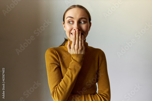 Image of cute funny girl of 20s having oops facial expression, covering mouth and looking aside with big round eyes after losing keys, making mistake at work, isolated on gray background photo